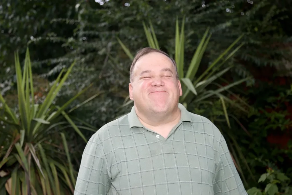 A man with a disability is smiling at the camera while outside in a garden